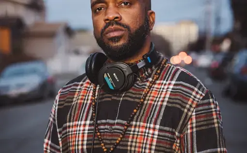 Headshot of Pendarvis Harshaw. He wears a plaid shirt and has headphones around his neck.