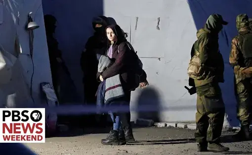 a refugee stands with blankets outside of a tent with soldiers
