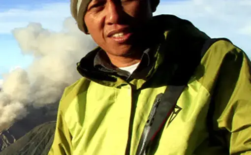 A man wearing a beanie and a yellow jacket. In the background, there is a mountain range and fog.