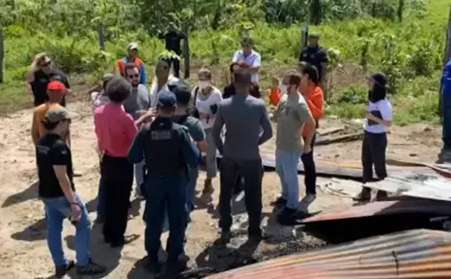 A crowd of people stand and discuss. In the foreground, there is a burned down building.