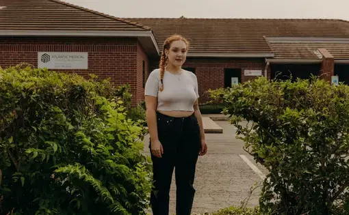 a young woman stands in front of a clinic that is overgrown with bushes and weeds