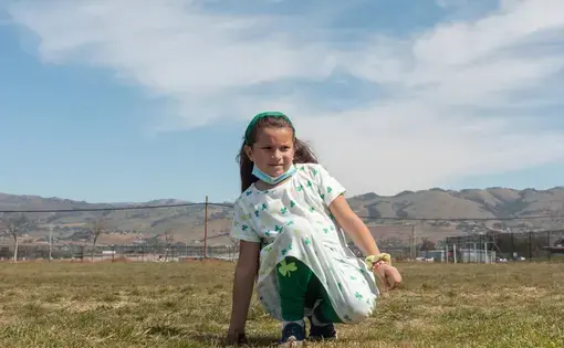 a girl kneels in a field, mountains in the background