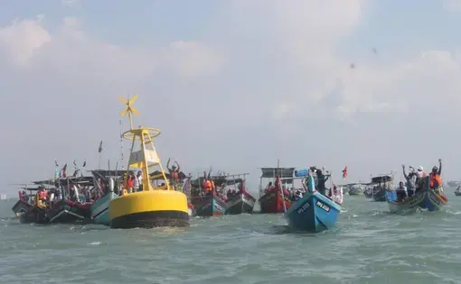 A series of small blue boats line up against the ocean. Fishers are waving hands and talking.