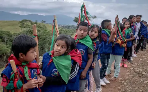 Children in blue vests and green-and-red bandanas tied around their necks line up on a mountain trail. Each carries a wood staff.
