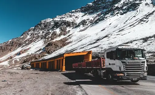 A large white truck drives through a yellow tunnel at the base of snow-dusted mountains.