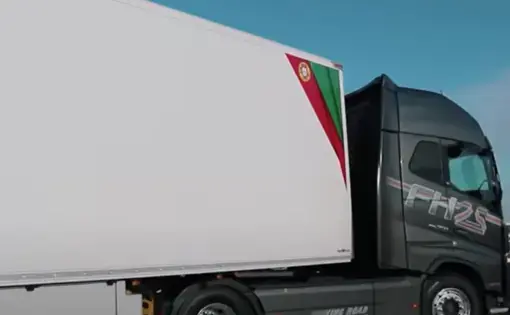 A grey truck turns with its red-and-green-striped, white payload in a parking lot