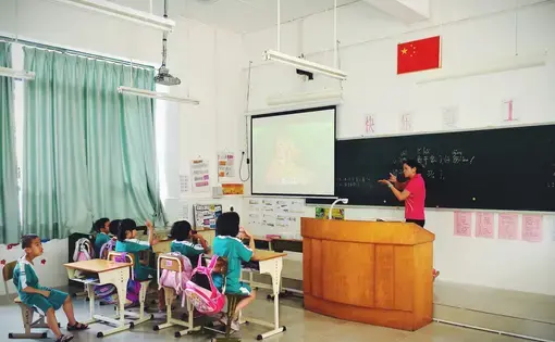 A teacher stands at the front of a classroom, gesturing toward the blackboard, while students raise their hands.