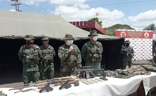 Men in military uniform stand behind a table laden with weapons. One man speaks into a microphone.