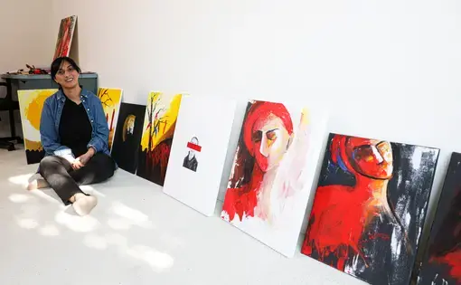 project page photo.jpg A woman sits on the floor next to a row of paintings that are on the floor propped up on a white wall