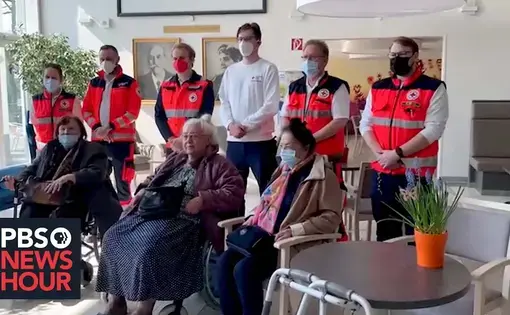 Six first responders in reflective orange uniforms featuring a red cross symbol stand behind three elderly women in wheelchairs.