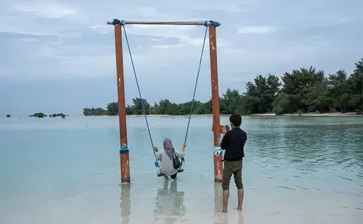 A woman swings on a submerged swingset while her companion takes her photo from behind. The swinging area is flooded by turquoise ocean water.