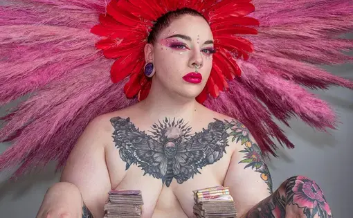 A shirtless woman with tatoos on her chest wearing a large, hot pink feather headdress sits at a table, where two large stacks of money cover her nipples.