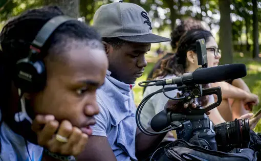 Four students work on creating a documentary. In the foreground, a student wears headphones to listen to audio they are recording. Another student uses a video camera to film. In the background, two students sit and watch the scene.
