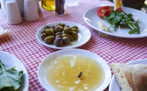 On a table covered in a tablecloth are dishes with olives, bread, vegetables, and honey, where a bee sits on the surface.