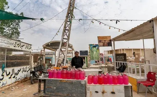 A man sells plastic jugs of a red-tinted liquid outside of a building.