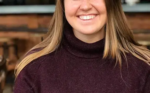 A woman with long blonde hair wearing a purple sweatshirt looks at the camera
