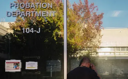 A person stands with their back facing the camera, making a call in front of a Probation Department window. A tree glows in the sunlight in the window's reflection.