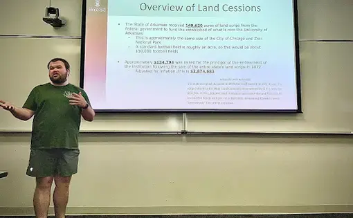 a young man in a green t-shirt and gray shorts stands in front of a class and presents a project in front of a projected presentation with the title "overview of land cessions"