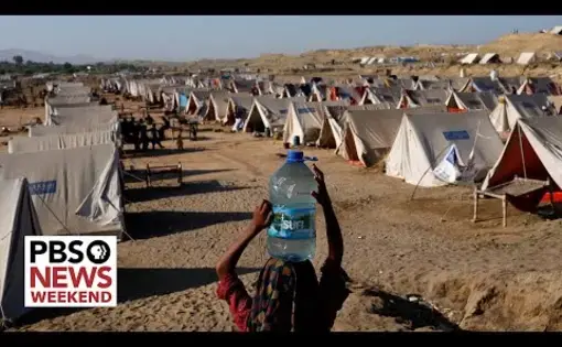 A person balances a plastic jug of water on their head while walking through an ailes of humanitarian tents