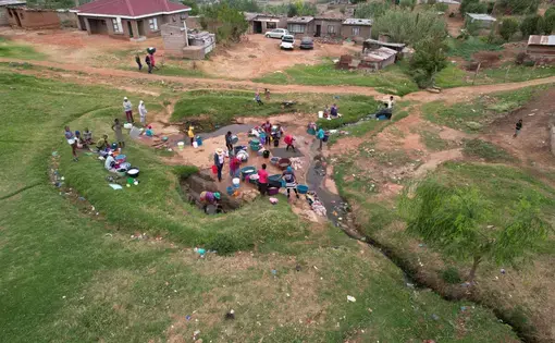 About thirty people gather around a small stream next to a dirt road. They carry washing bins and some are bending over the water.