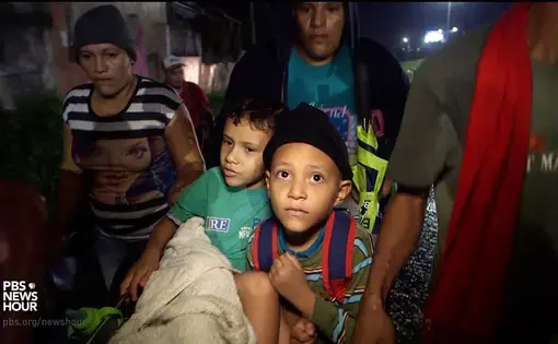 Two young boys sit on a cart being pushed at night by three adults.