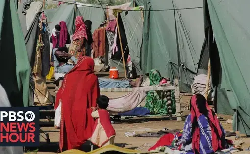 Youtube thumbnail for the PBS Newshour report. Women in colorful robes tend to children and sit in a camp lined with makeshift green tents.