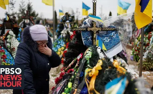 A woman in winter clothes cries as she stands next to a heavily decorated gravesite in a field of similar gravesites, decorated with flowers, ribbons with messages, crucifixes, and the Ukrainian flag.