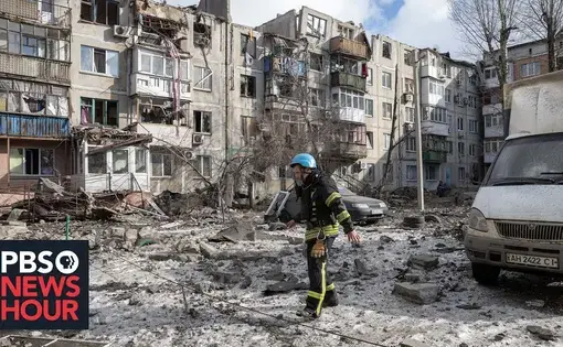 A man in firefighter gear and helmet traverses a plaza littered with debris from destroyed apartment buildings.