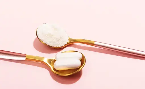 Collagen powder and pills in two gilded, parallel spoons on a pink background.