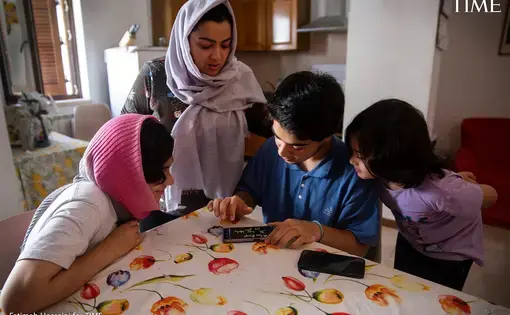 A woman wearing a headscarf sits with three children at a kitchen table. They all look at a cell phone message