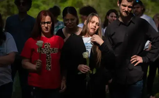 A reflection iluminates the face of the mother of a deceased youth. She and others are holding white roses. Her T shirt reads 'justice for Joshua Beasley.'