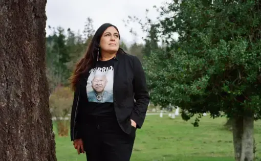 A woman stands in a cemetery near a tree. She wears a T shirt with the image of an elder and the words "Warrior"