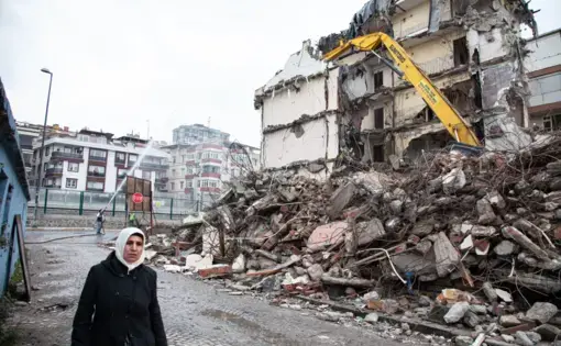 A woman walks down a street past a dilapidated apartment structure, where heavy machinery is at work and workers spray water on the demolition site.