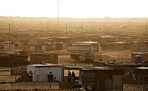 The sun sets over a settlement composed of makeshift homes and numerous streetlight poles.