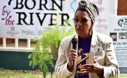 A woman talks into a microphone in front of a mural that reads "Born by the River."