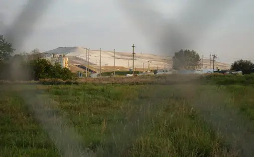 A mountanous pile of sand in an empty field. The image is taken through a chainlink fence surrounding the mine.