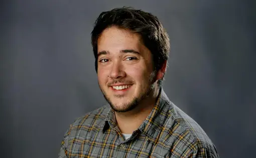 A man with short dark hair wears a plaid button down against a dark background