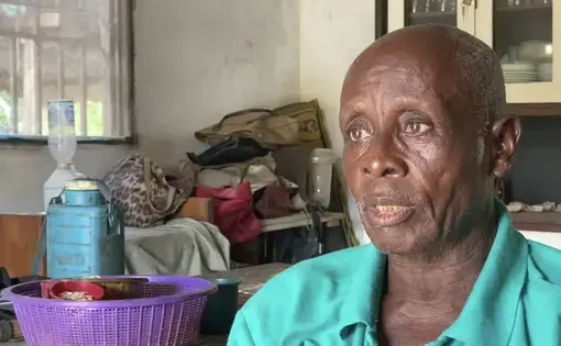 An older man wearing a blue shirt is interviewed in his home