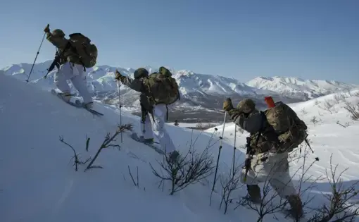 Three individuals in hiking gear climb a snowy ridge with high mountains in the background.