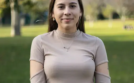 A woman with long dark hair stands against a grassy background.