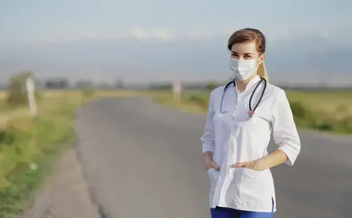 a woman wearing a white medical coat and a face mask stands on an empty road with flat green expanses behind her