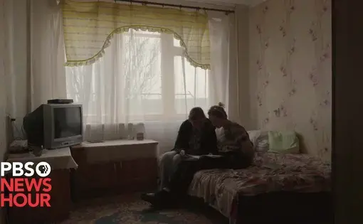 A mother and child sit on the bed in a dark bedroom, looking at a book.