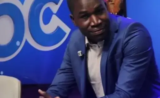 A man sits in a sharp blue suit with his hands folded.