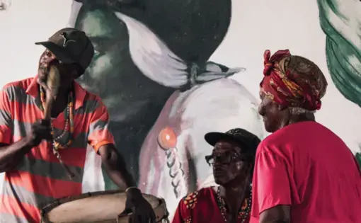 Three people play drums in front of a mural in Jamaica
