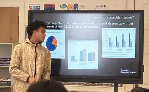A teen stands in front of a screen and presents his presentation on immigration by displaying a slide with several graphs