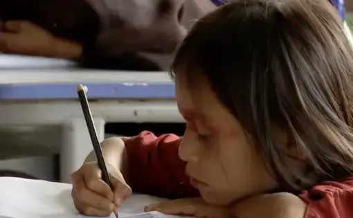 A little girl in red sits with her head resting on the desk, writing with a pencil.