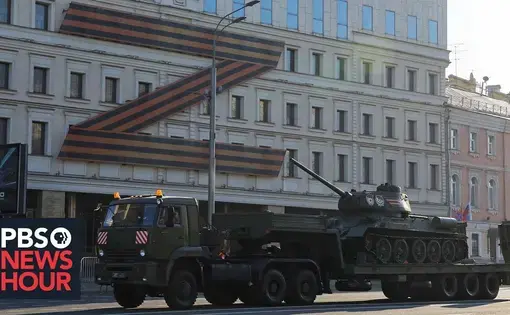 A tractor trailer carrying a tank is parked outside of a building marked with a large black Z logo