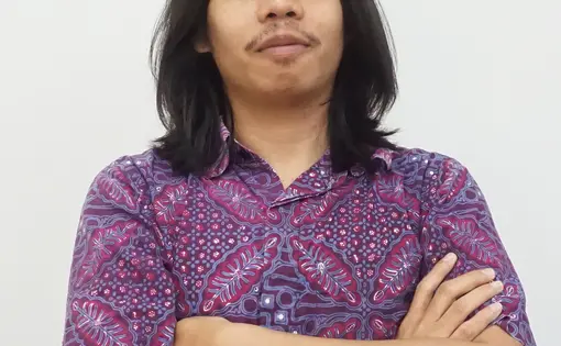 A man with long dark hair and wearing a purple shirt folds his arms