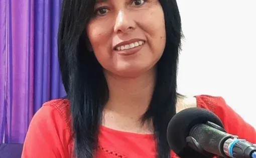 A woman with long dark hair wears a pink dress and speaks into a microphone