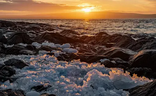 The sun setting over the sea, with broken-up ice along a rocky shoreline in the foreground